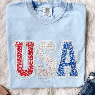Chambray Comfort Colors tee folded flat with floral patterned USA letters in red white and blue with lace border detail
