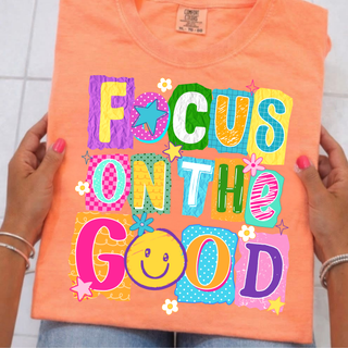 Hand holding neon cantaloupe tee with FOCUS ON THE GOOD colorful daisy pop patchwork letters, smiley face, youth adult unisex