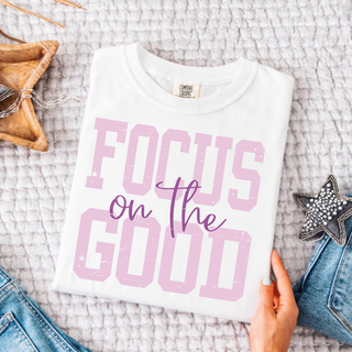 White Comfort Colors tee with pink and purple Focus on the Good graphic held against neutral background