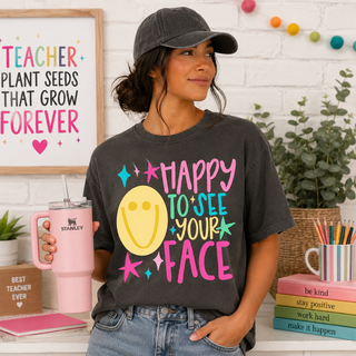 pepper grey adult graphic tee with happy to see your face teacher design, smiley face, model at desk with pink stanley cup
