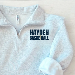 Close-up of Hayden Basketball lettering with a cross mark printed on the chest of a cadet collar quarter-zip.