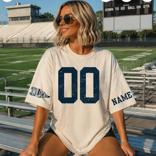 Sand oversized tee with distressed navy custom number on the chest, a Wildcats pennant flag icon on one sleeve, and a personalized name on the other sleeve