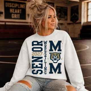 Woman sitting in a wrestling room wearing a light grey crewneck sweatshirt with vertical Senior 2027 Wrestling lettering beside a navy M, gold and navy wildcat head, and second M