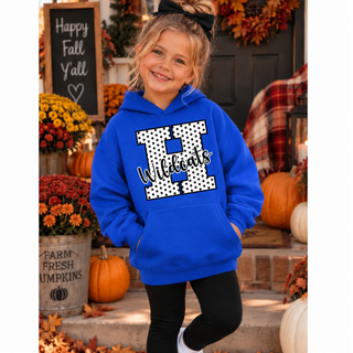 Girl in a royal blue hoodie with a white polka dot letter H outlined in black and black Wildcats script on the front, with plain sleeves, standing on a fall porch with pumpkins
