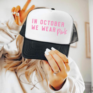 Black and white trucker hat with pink In October We Wear Pink lettering and script design
