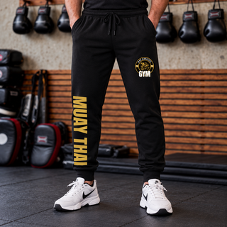 Man in black core fleece joggers in a gym with the Iron Warriors Gym lion crest on the upper leg and Muay Thai in gold vertical lettering down the leg