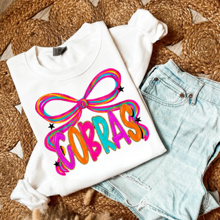 White Jackson Heights Cobras Bow Top folded flat, featuring a colorful rainbow bow with bold multicolor COBRAS lettering and star accents