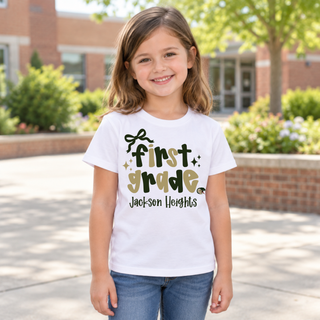 Girl wearing Jackson Heights Cobras First Grade Bow Top in white featuring first grade bubble lettering with a bow accent, sparkles, Jackson Heights script, and a cobra head icon