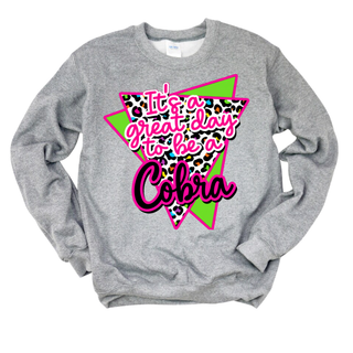 Grey Jackson Heights Cobras Great Day Top folded flat, featuring "It's a great day to be a Cobra" script lettering over a leopard print pattern inside a neon green and hot-pink retro triangle graphic