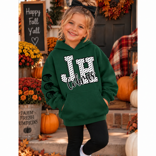 Smiling girl in a forest green hoodie with a white polka dot JH outlined in black, black Cobras script, and three cobra head logos down the sleeve, standing on a porch with pumpkins and mums