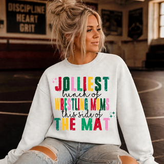 Woman in a white crewneck sweatshirt with the Jolliest Bunch of Wrestling Moms This Side of the Mat graphic in red, green, pink and yellow block letters with black script, sitting in a wrestling room