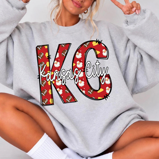 Woman in an oversized ash grey sweatshirt with red doodle KC letters filled with footballs and Santa faces and white script Kansas City lettering
