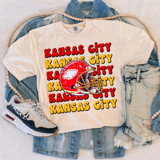 Cream tee with Kansas City repeated in stacked red and gold lettering and a red football helmet graphic, flat lay with denim jacket and shorts
