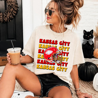 Cream tee with Kansas City repeated in stacked red and gold lettering and a red football helmet graphic, worn by a model