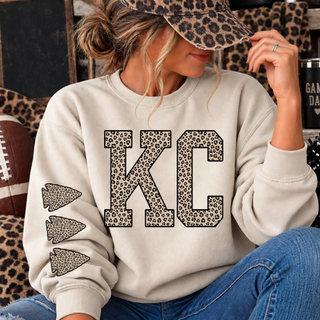 Cream crewneck sweatshirt with KC lettering in leopard print and three leopard print arrowheads down the sleeve, worn by a model