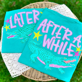 Island green Comfort Colors tees with Later Alligator After a While Crocodile alligator graphic and pink and yellow star design folded in basket