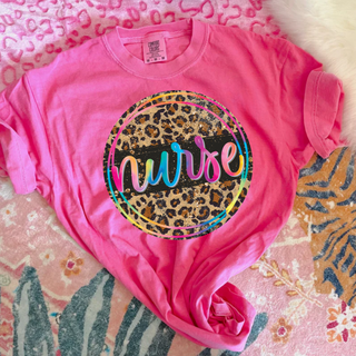 Neon pink tee laid flat showing a leopard print circle graphic with colorful rainbow nurse script in the center
