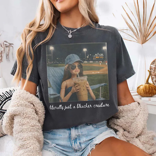 Woman wearing a Comfort Colors Pepper tee with an AI generated photo style image of a girl at a baseball game holding an iced coffee with the words literally just a bleacher creature