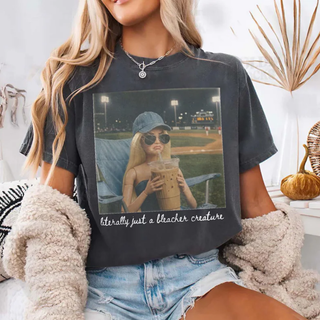 Close up of Comfort Colors Pepper tee with an AI generated photo style image of a girl in sunglasses and a baseball cap at a night game holding iced coffee with the words literally just a bleacher creature