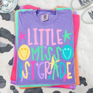 Little Miss 1st Grade graphic tee in violet folded with colorful lettering, stars, and smiley faces
