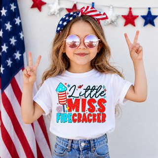 Little girl wearing white tee with little miss firecracker red white blue rocket design sunglasses flag bow with peace signs

