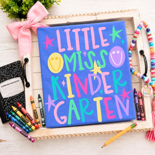 Little Miss Kindergarten graphic tee in flo blue with colorful lettering, stars, and smiley faces on styled tray
