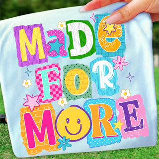 Hand holding chambray tee with MADE FOR MORE colorful daisy pop patchwork letters, smiley face, and flowers