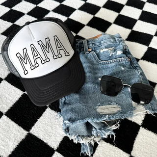 Black and white foam front trucker hat with bold outlined MAMA varsity lettering styled with denim shorts and sunglasses on a checkered rug