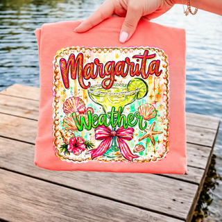 Hand holding a coral tee by the water with Margarita Weather glitter script margarita glass bow and tropical graphic
