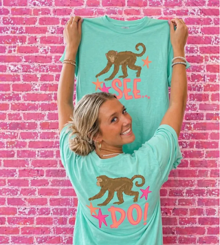 Woman holding up chalky mint Monkey See Monkey Do Graphic Tee showing front SEE and back DO monkey design with pink and orange stars