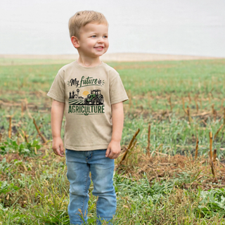 Toddler boy wearing tan My Future is Agriculture tee with tractor and farm graphic in a field