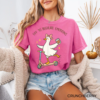 Woman wearing a Comfort Colors Crunchberry tee with a cartoon goose riding a scooter holding coffee and the words off to regulate emotions