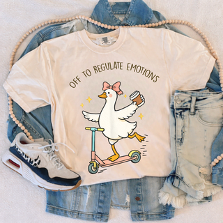 Ivory Comfort Colors tee with a cartoon goose riding a scooter holding coffee and the words off to regulate emotions laid flat with denim jacket and sneakers