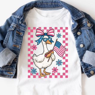 White toddler graphic top with patriotic goose wearing bow and sunglasses holding American flag on pink checkered background with denim jacket