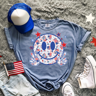 Comfort Colors blue jean short sleeve top laid flat with red white and blue floral American flag smiley face peace sign graphic styled with denim shorts and white sneakers
