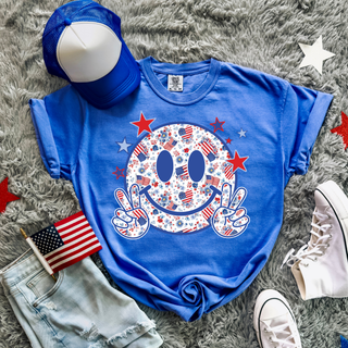 Comfort Colors flo blue short sleeve top laid flat with red white and blue floral American flag smiley face peace sign graphic styled with denim shorts and white sneakers
