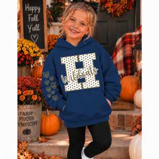 Young girl in a navy hoodie with a white polka dot H outlined in gold, Wildcats in gold script, and gold wildcat faces down the sleeve, standing on a fall porch with pumpkins and mums