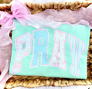 Pray graphic top in pastel blue, pink, and floral patterned letters on mint green background, folded in woven basket with pink ribbon