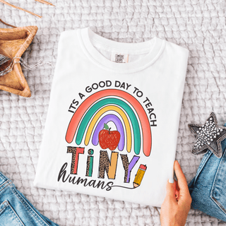 White Comfort Colors tee folded flat with it's a good day to teach TINY humans rainbow apple and leopard print design
