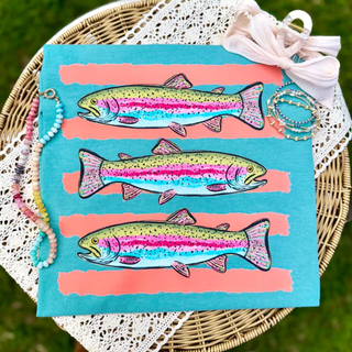 Teal tee laid flat on wicker basket showing three stacked colorful rainbow trout fish on coral stripe graphic
