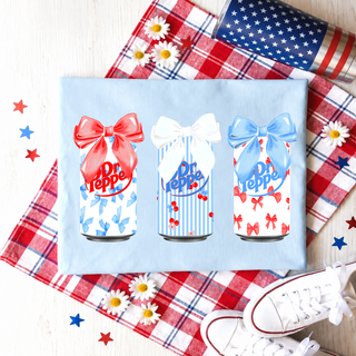 Red White and Blue Soda Bow Graphic Top in light blue with three soda cans with red white and blue bows flat lay styled with patriotic decor
