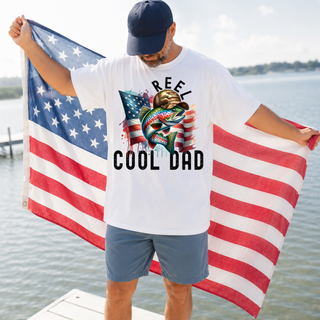 Man wearing Reel Cool Dad Shirt white tee with rainbow trout, camo hat, American flag graphic holding American flag on dock