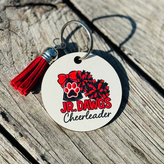 Round white keychain with JR. DAWGS Cheerleader megaphone paw print pom pom design and red tassel on wood background