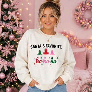 Model wearing the Santa's Favorite Ho Ho Ho top in cream with black Santa's Favorite lettering, three Christmas trees, and script ho ho ho against a pink Christmas backdrop