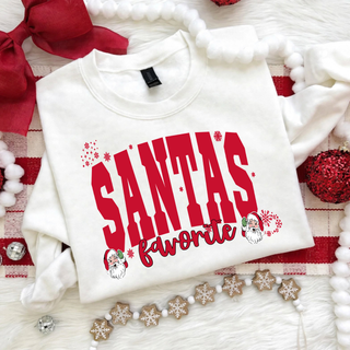 Santa's Favorite design in red arched block lettering with a script favorite and two retro Santas with snowflakes on a white sweatshirt, flat lay with red bow and pom pom holiday decor