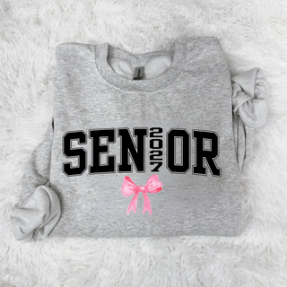 Flat lay of a heather grey crewneck sweatshirt with black varsity SENIOR lettering, a vertical 2027 as the I, and a pink watercolor bow, on a white faux fur rug