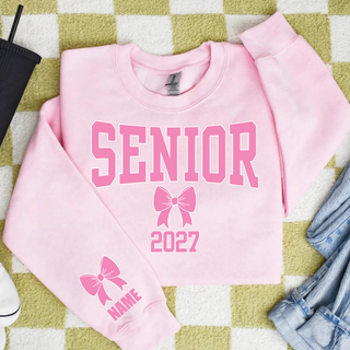Flat lay of a light pink Gildan crewneck sweatshirt with pink varsity SENIOR lettering, a pink bow, and 2027, plus a pink bow and NAME on the sleeve, on a green checkered rug