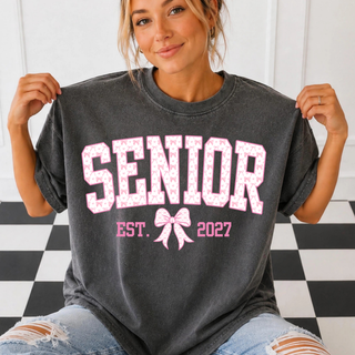 Woman holding out a pepper gray Comfort Colors tee with varsity SENIOR letters in a pink bow and heart pattern, a pink bow, and EST. 2027