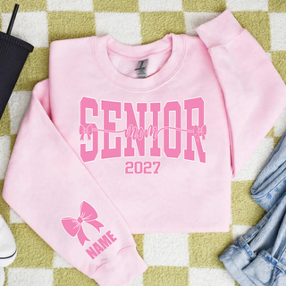 Flat lay of a light pink crewneck sweatshirt with pink varsity SENIOR lettering, script mom with small bows, 2027, and a pink bow with NAME on the lower sleeve
