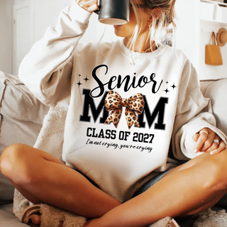 Woman in a white crewneck sweatshirt sipping from a mug, with a Senior Mom Class of 2027 design featuring black varsity MOM letters, a leopard print bow, sparkles, and the words I'm not crying, you're crying